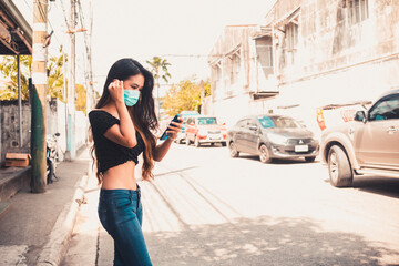 A slim Filipina woman in a crop top uses a ride sharing app and waits for a car while wearing a face mask to shield from pollution. In a typical Manila neighnorhood. - obrazy, fototapety, plakaty