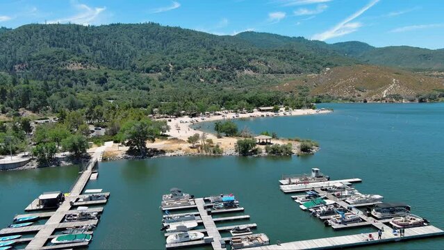 aerial footage of the rippling blue waters of Silverwood Lake with boats docked in the marina, a beach with people relaxing, mountains covered with lush green trees and plants in Hesperia California