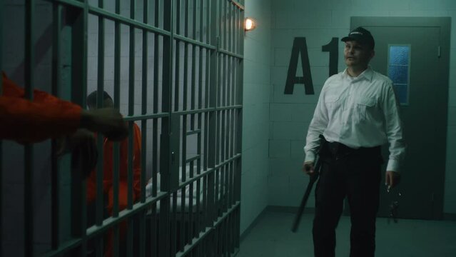 Warden with baton and bunch of keys comes to jail cells and talks with prisoners. African American men in orange uniform serve imprisonment term in prison cells. Guilty murderers in detention center.