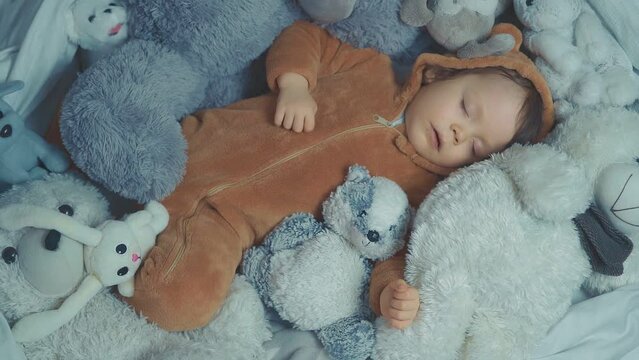 Little baby with outstretched hands lying in bed with fluffy stuffed toys of animals. Toddler sleeping with teddy bear in cozy room and enjoy healthy night sleep. Selective focus. Top view.