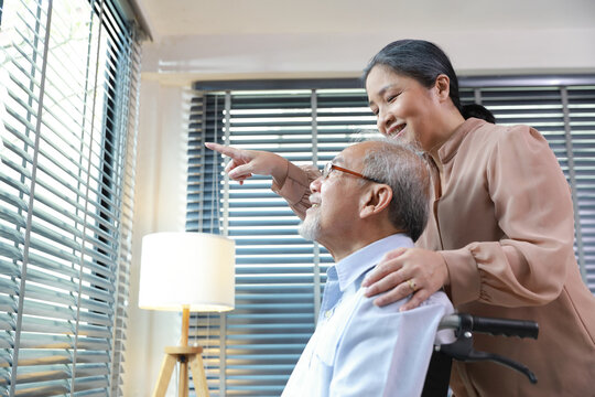 Asian Senior Woman Or Caregiver Helping And Consoling Senior Man Walk With Wheelchair In Living Room. Elderly Wife Taking Good Help Care And Support Of Elder Husband Patient Inside The House.