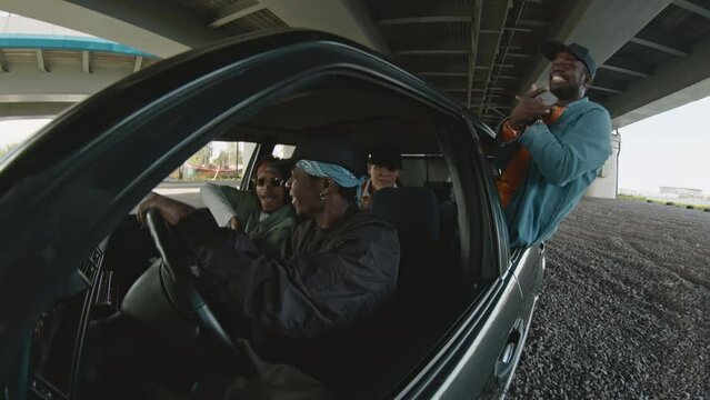Four young diverse men wearing oversized clothes, bandanas and caps listening and dancing to hip hop and rap music in black retro car with open windows