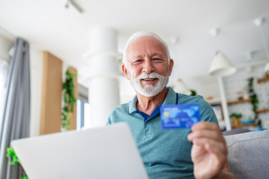Mature man using laptop, holding plastic credit or debit card, senior grey haired customer making secure internet payment, shopping or browsing online banking service, entering information