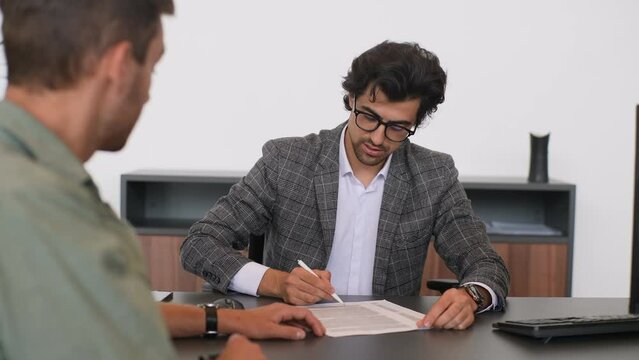 Portrait of male real estate broker wearing business suit present and advise young man client on decision to sign insurance contracts. Happy male customer signing purchase agreement and shaking hands.