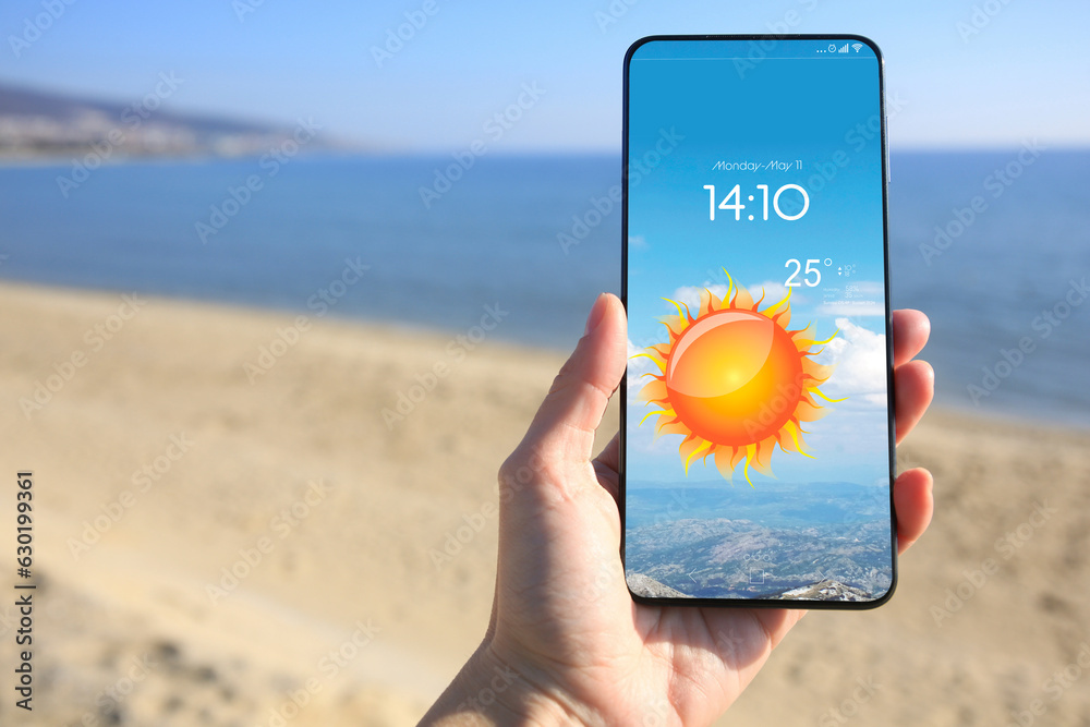 Wall mural man checking weather using app on smartphone on beach, closeup. data and illustration of sun on scre