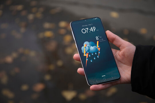 Woman checking weather using app on smartphone on street, closeup. Data and illustration of cloud with lighting and rain on screen