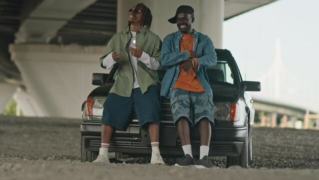Two African American mates listening and dancing to hip hop music while leaning on hood of their old school car parked under bridge