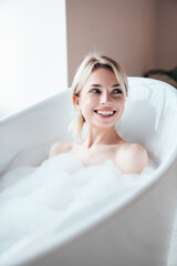 Fototapeta na wymiar Portrait of young beautiful sexy woman having fun while lying in bathtub full of foam at home. Charming smiling model relaxing in luxury bath interior. Female enjoying beauty and skincare day