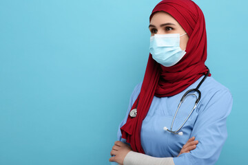 Muslim woman wearing hijab, medical uniform and protective mask on light blue background, space for...