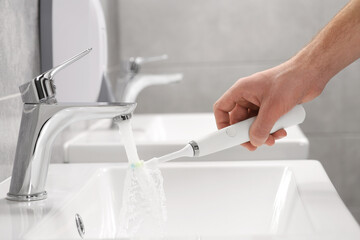Man holding electric toothbrush under flowing water above sink in bathroom, closeup