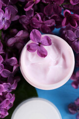 Obraz na płótnie Canvas Jar of cream and lilac flowers on light blue table, flat lay