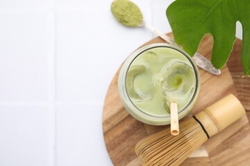 Glass of tasty iced matcha latte, leaf, bamboo whisk and powder on white tiled table, flat lay. Space for text