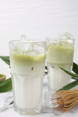 Glasses of tasty iced matcha latte, bamboo whisk and leaves on white marble table