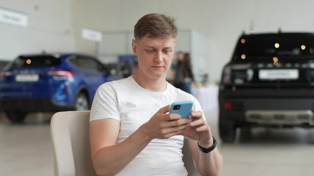 Portrait of handsome focused customer male using smartphone sitting on armchair at office table in dealership store office. Young man client buyer choose auto want buy new car at showroom, slow motion