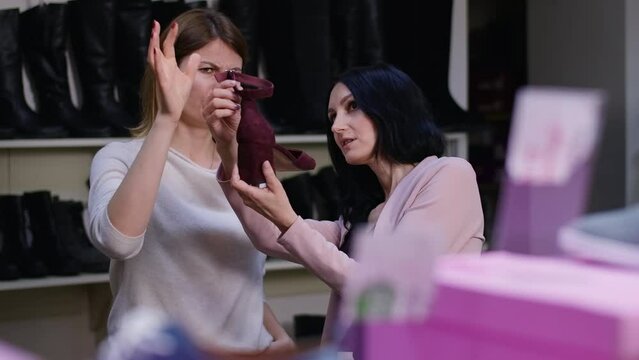 Shop assistant offering new high-heels to unsatisfied client with dissatisfied facial expression. Portrait of picky Caucasian woman making decision choosing footwear with help of employee in shop