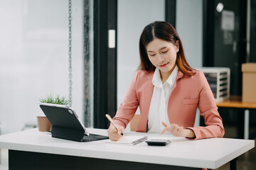 Young beautiful Asian woman typing on tablet and laptop while sitting at the working white table modern office