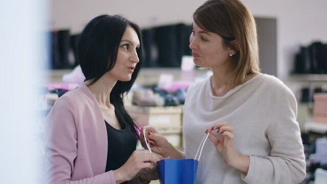 Satisfied woman bragging new shoes to friend standing in shoe shop indoors. Smiling happy Caucasian lady with shopping bag showing off on Black Friday
