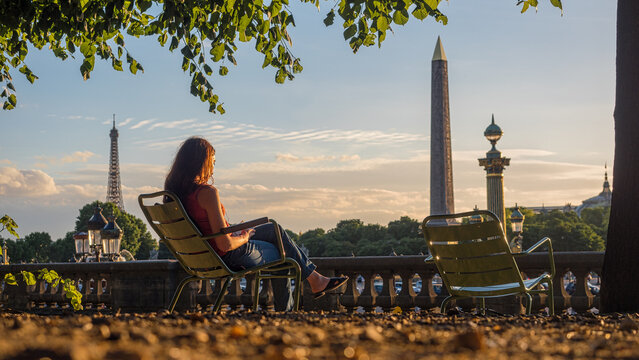 Jeune femme assise sur un chaise publique du Jardin des Tuileries lit un livre au soleil, vue sur Tour Eiffel et l'Ob&eacute;lisque de la place de la Concorde