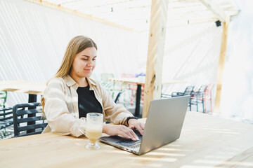 Fototapeta na wymiar Young happy woman sitting at outdoor cafe table and talking on phone with cup of coffee, smiling woman enjoying telecommuting in cafe or studying online on laptop
