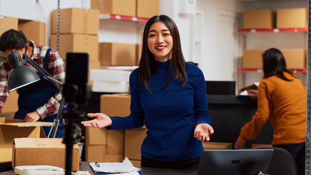 Business owner filming teleshopping video on camera, advertising merchandise in storage room. Young woman using social media streaming platform to create online promotion, marketing. Tripod shot.