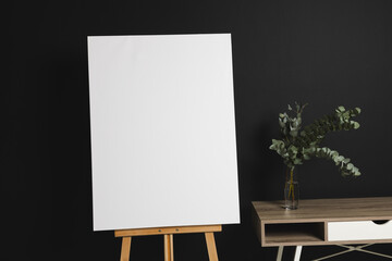 White canvas on wooden easel and copy space with desk and plant on black background