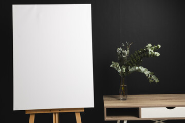White canvas on wooden easel and copy space with desk and plant on black background