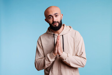 Hopeful arab man looking at camera with folded hands in pleading gesture. Young handsome person wearing casual hoodie praying, making praise sign with arms studio portrait