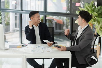 Excited happy young Asian two businessman winner and celebrating success at desk in office.