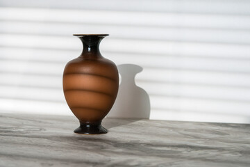 Brown vase on a gray marble tabletop near a white wall illuminated by sunbeams through the blinds