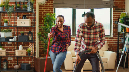 Fototapeta na wymiar Happy man and woman showing dance moves and doing spring cleaning, having fun with music cleaning floors with mop and vacuum cleaner. Young smiling couple doing chores. Handheld shot.