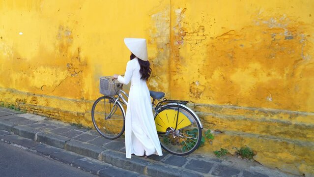 Asian woman wearing white Vietnamese culture with traditional conical hat with bicycle in Hoi An, Vietnam.