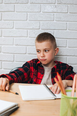 Concept back to the school. An upset boy in the process of studying sits at a table with books, notebooks and pencils. Front view