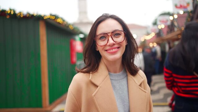 Portrait shoot elegant young woman in beige coat and horn-rimmed glasses outside warm winter day. Beautiful female in good mood standing european Christmas market, looking camera and smiling