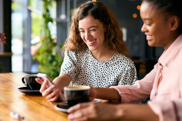 Two Young Female Friends Meeting In Coffee Shop And Looking At Mobile Phone Together