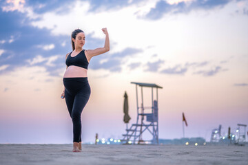 Fototapeta na wymiar Nine months pregnant woman in sportswear showing muscles, standing on the sandy beach at sunset. Healthy lifestyle, sport and pregnancy concept.