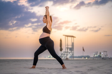 Side view of pregnant woman in black sportswear making Crescent Lunge yoga pose on the beach at...