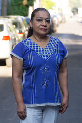 Mature Mexican lady looking at the camera smiling,wearing a traditional mexican embroidered blouse standing, with out of focus background of street. Latin woman looking at camera