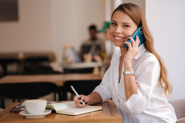 Financial manager writing down in notebook and speak by phone in same time. Working in cafe. Freelancer concept