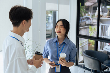 two young asian businesswomen standing chatting talking and holding coffee by the window in office