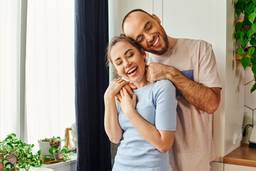 Joyful bearded man in homewear embracing girlfriend with closed eyes while standing at home