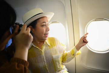 Smiling middle aged woman and daughter sitting in passenger airplane and taking picture, waiting for airplane landing