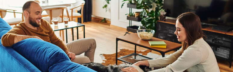 Obraz premium Smiling woman petting border collie dog on couch near boyfriend in living room, banner