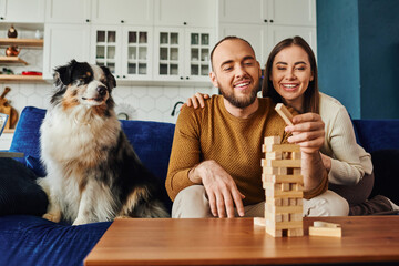 Smiling woman hugging boyfriend and playing wood blocks game near border collie dog on couch at home