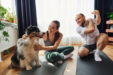 Smiling man warming up near girlfriend in sportswear petting border collie on fitness mat at home