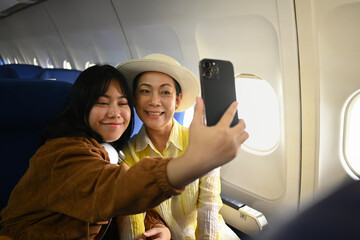 Smiling middle aged woman and daughter sitting in passenger airplane and taking picture, waiting...