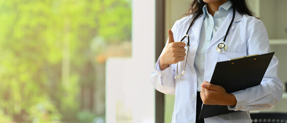 Doctor dressed in white overall with stethoscope holding clipboard and showing thumbs up