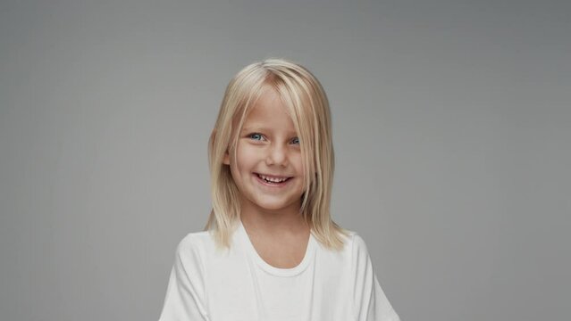 Portrait of Young Girl with Smiling Face Looking at Camera in Colour Studio Shot. Happy Cheerful Kid Isolated Alone on Grey Background Closeup. Emotional Child with Bright Blue Eyes in White Clothes