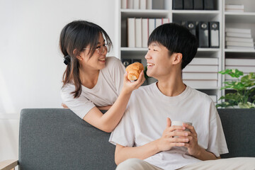 Young couples spend holidays in the living room. The young man was wearing comfortable clothes, showing his face, satisfied with the taste of snacks. Eating croissant on the sofa