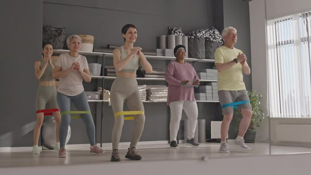 Full shot of group of elderly people doing resistance band side steps in front of mirror while exercising in gym under control of female fitness coach