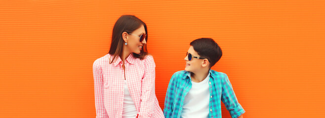 Stylish happy smiling mother with son teenager posing together in sunglasses, checkered shirts, jeans in the city on orange background
